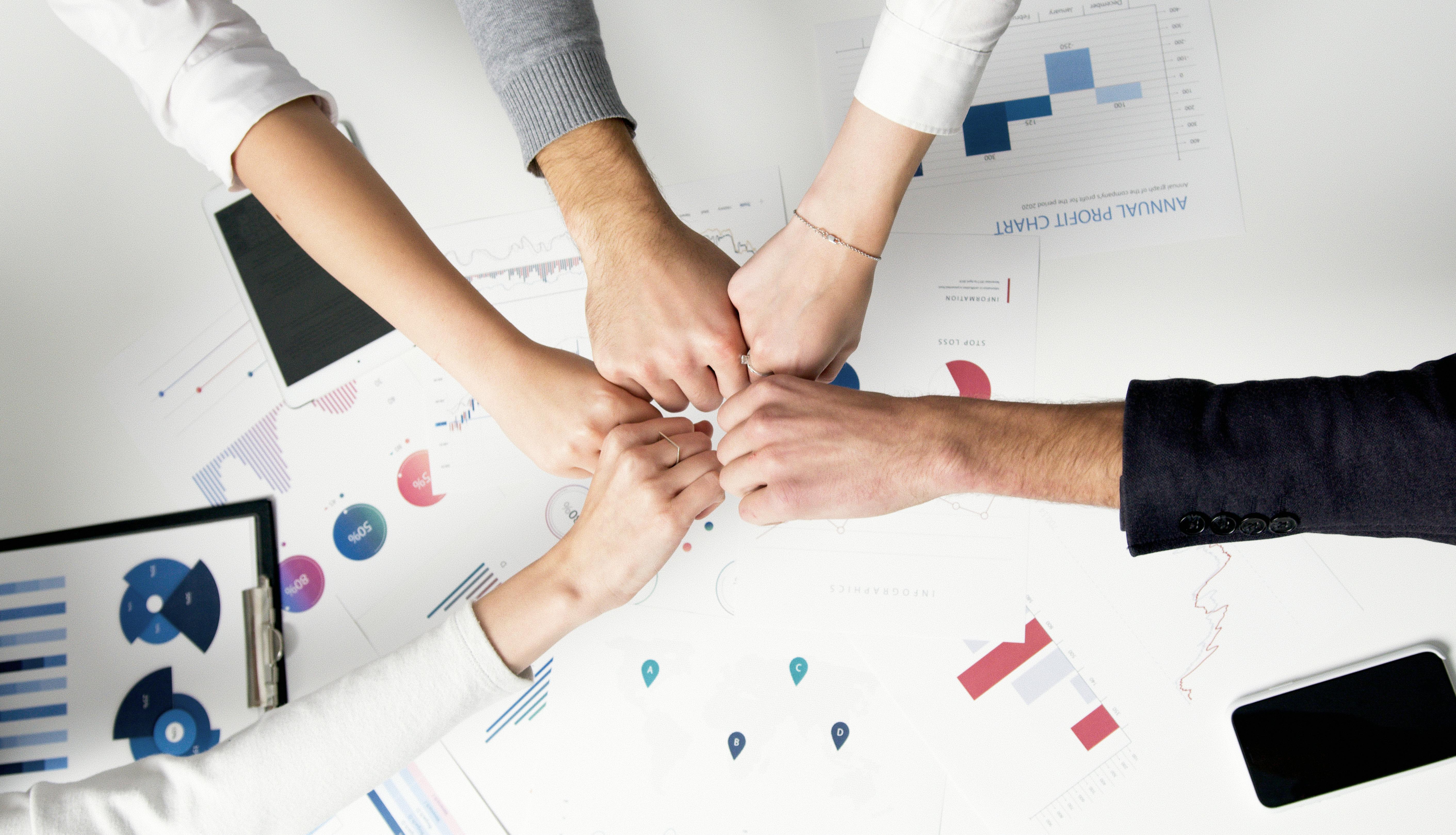 5 people, with only their hands and arms showing, fist- umping each other above a table full of papers and a clipboard