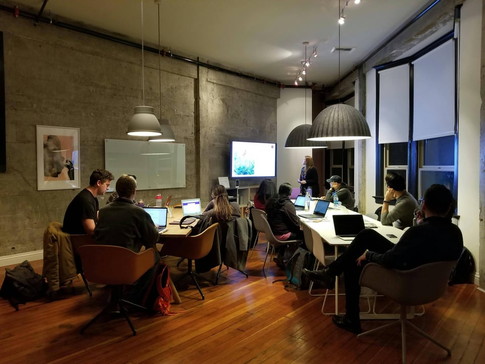 group of people on their laptops with a woman standing in front of them discussing using the TV