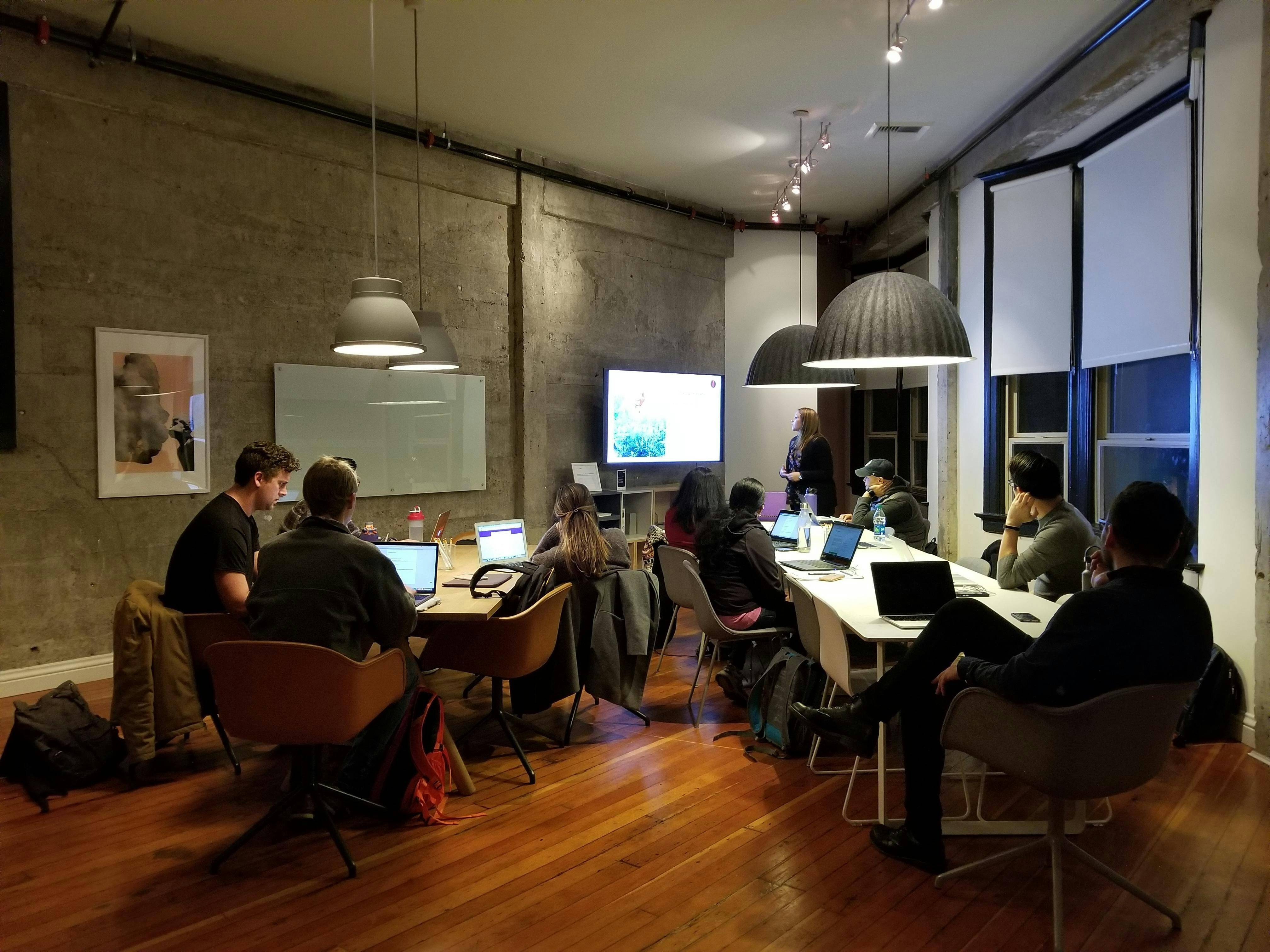 group of people in front of their laptops with a woman in front discussing in front of a TV